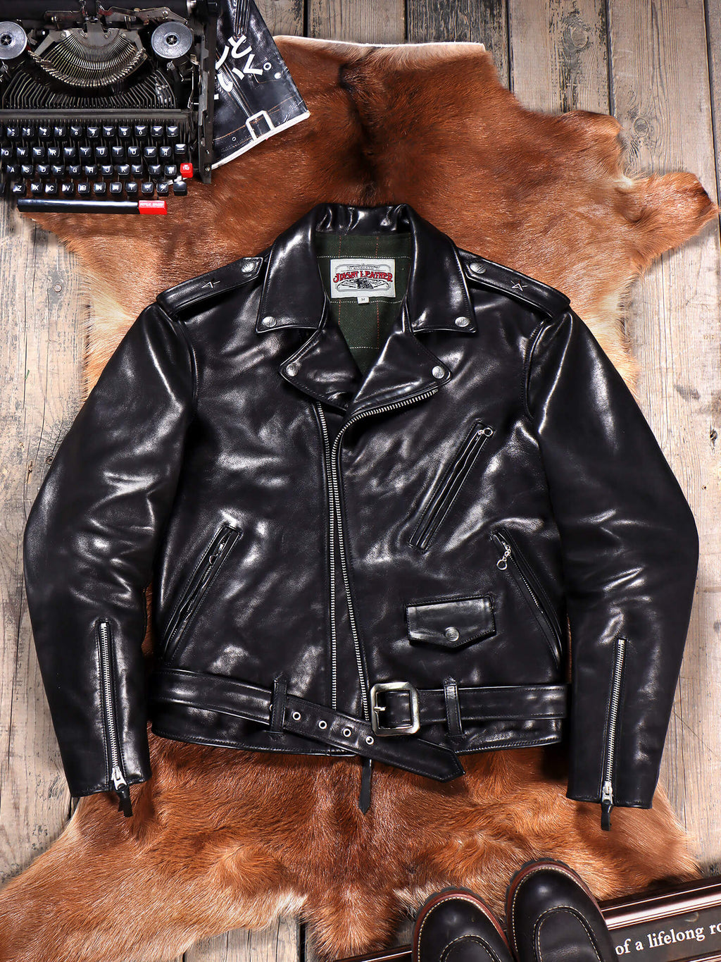 A black leather motorcycle jacket with a belted waist and zippered pockets, laid flat on a brown animal hide over a rustic wood floor. A vintage typewriter appears in the top-left corner and dark shoes are partially visible at the bottom.