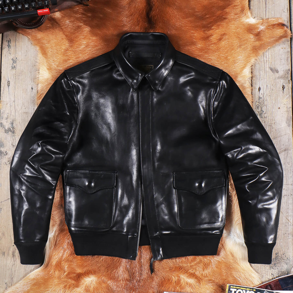 A black leather bomber jacket with a front zipper, ribbed collar, cuffs, and hem, laid flat on a brown animal hide over a rustic wooden surface.