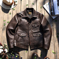 A dark brown leather jacket laid flat on a rustic wooden surface, surrounded by styled props including a hat, flowers, a book, and boots. The jacket has a pointed collar, button front, and four front flap pockets with metal snap buttons.