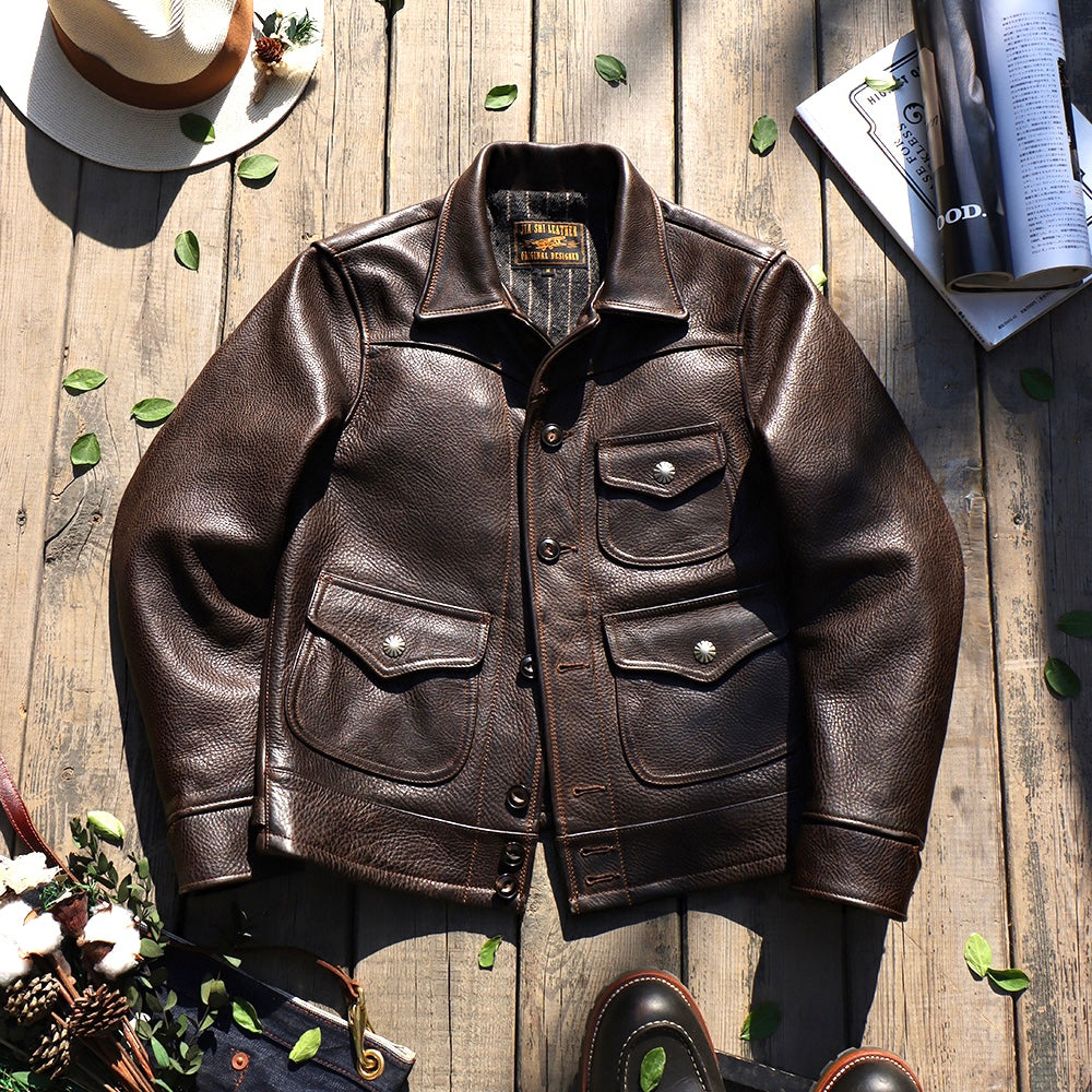 A dark brown leather jacket laid flat on a rustic wooden surface, surrounded by styled props including a hat, flowers, a book, and boots. The jacket has a pointed collar, button front, and four front flap pockets with metal snap buttons.