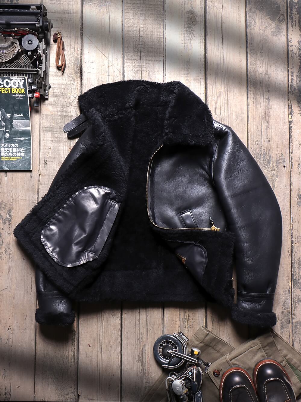 A black leather and shearling jacket laid flat on a rustic wooden surface, styled with vintage props including a typewriter, a book, and shoes.