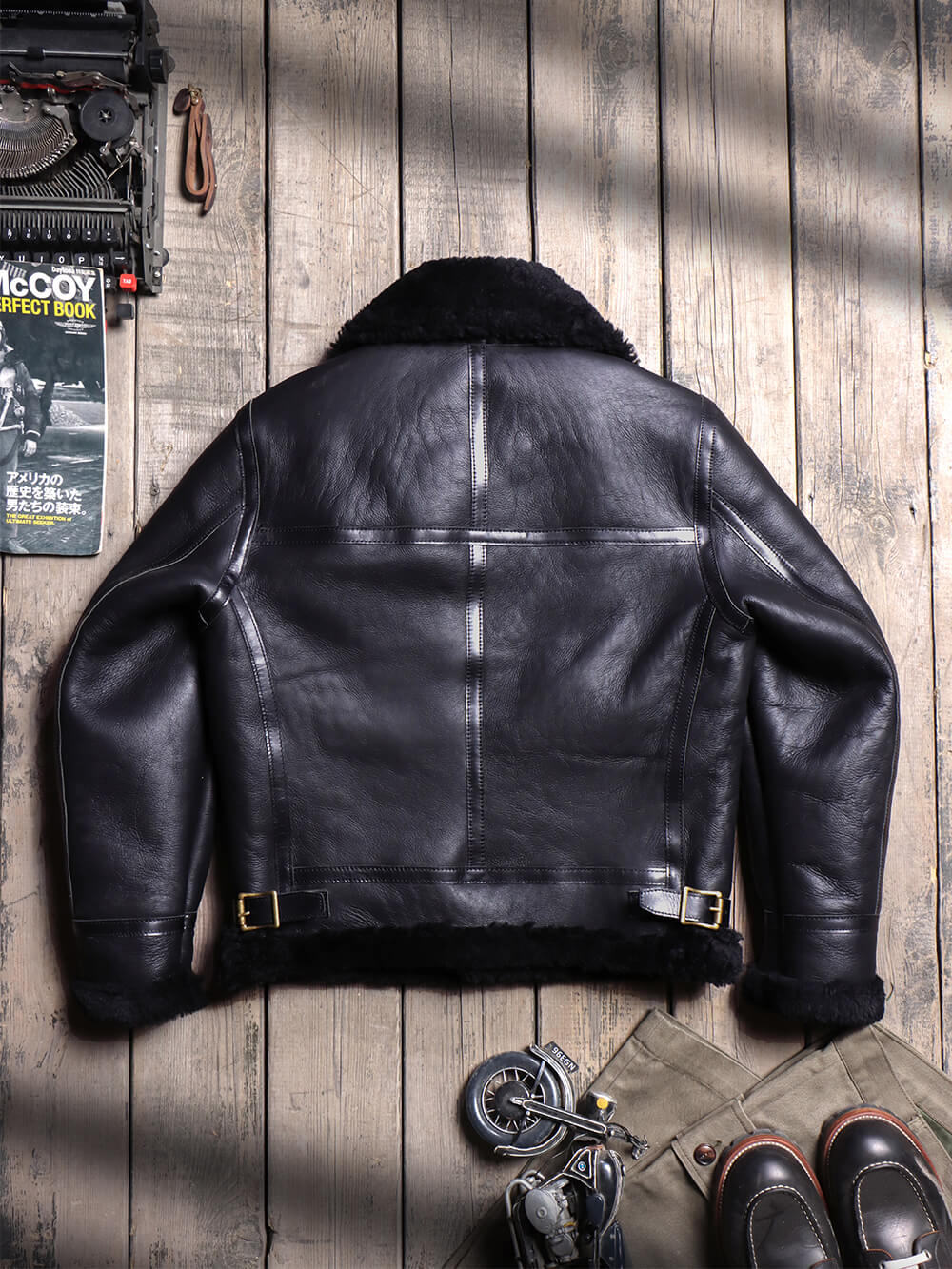A black leather aviator-style jacket with a dark shearling collar, laid flat on a rustic wooden plank background. The jacket is shown from the back, featuring a center seam, horizontal chest paneling, and a belted waist with brass-tone hardware.